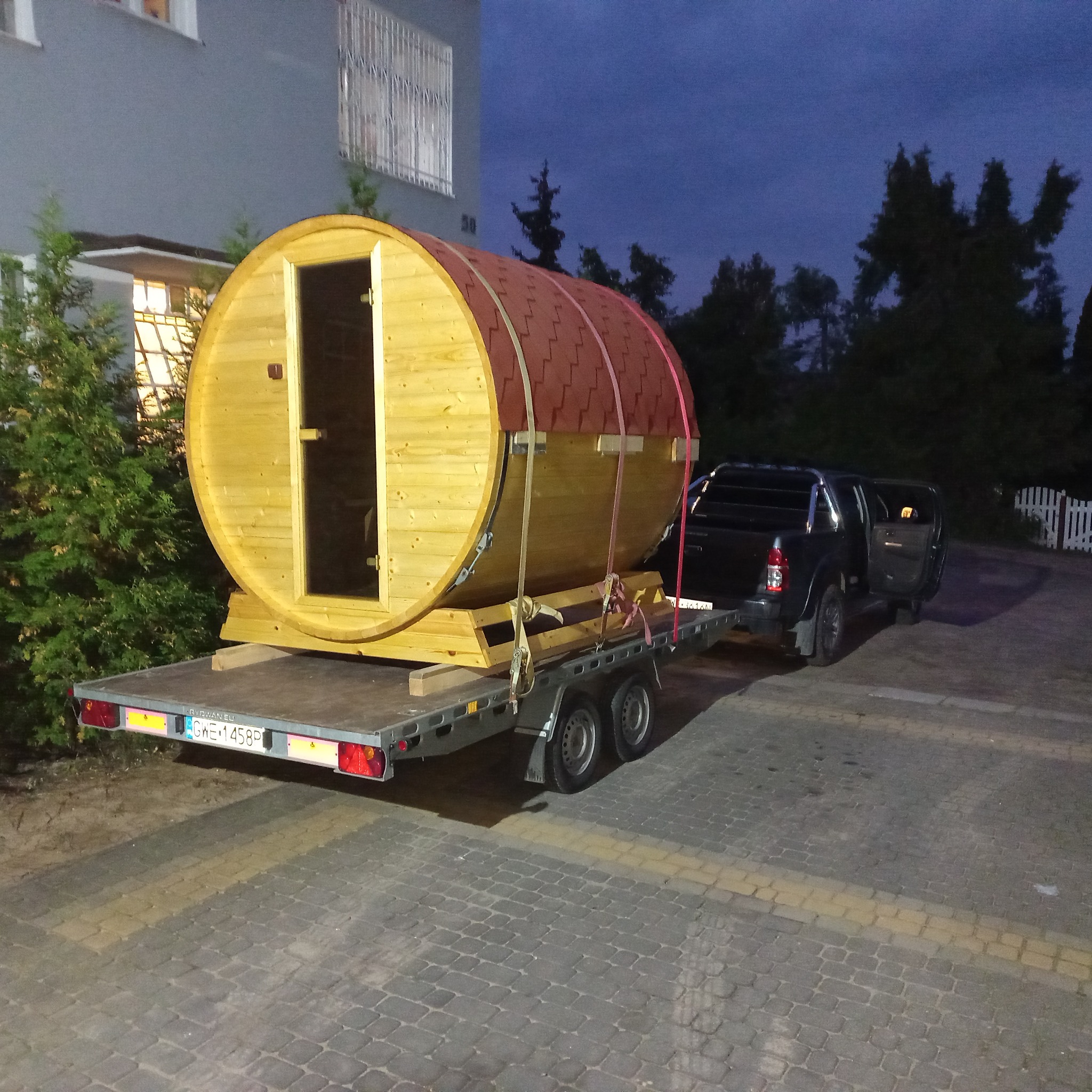 Drewniana beczka z drzwiami, imitująca saunę, zabezpieczona pasami na przyczepie samochodowej podczas transportu, na tle budynku mieszkalnego i drzew.