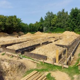 SETA-BUD sp. z o.o. - Przygotowanie fundamentów pod budynek: widoczne zbrojenia w szalunkach, ziemia usypana w kopce, w tle budynek w trakcie budowy.