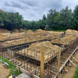 SETA-BUD sp. z o.o. - Zbrojenie fundamentów domu jednorodzinnego, widoczne szalunki, pręty stalowe i częściowo zasypany wykop ziemią, w tle budynek w stanie surowym otoczony zielenią.