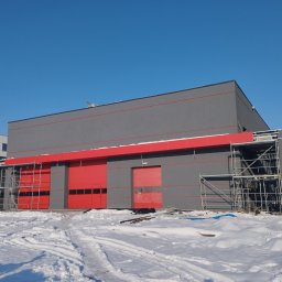 SETA-BUD sp. z o.o. - Zewnętrzny widok nowo wybudowanego, szarego budynku z czerwonymi bramami garażowymi, otoczonego śniegiem i rusztowaniami.