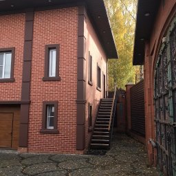 SETA-BUD sp. z o.o. - Dwupiętrowy dom z czerwonej cegły z brązowymi detalami w Warszawie, widok na schody zewnętrzne i ozdobną bramę wjazdową z boku budynku.