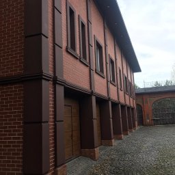 SETA-BUD sp. z o.o. - Budynek z czerwonej cegły z brązowymi elementami dekoracyjnymi, widoczne okna i brama garażowa, brukowana droga z liśćmi.