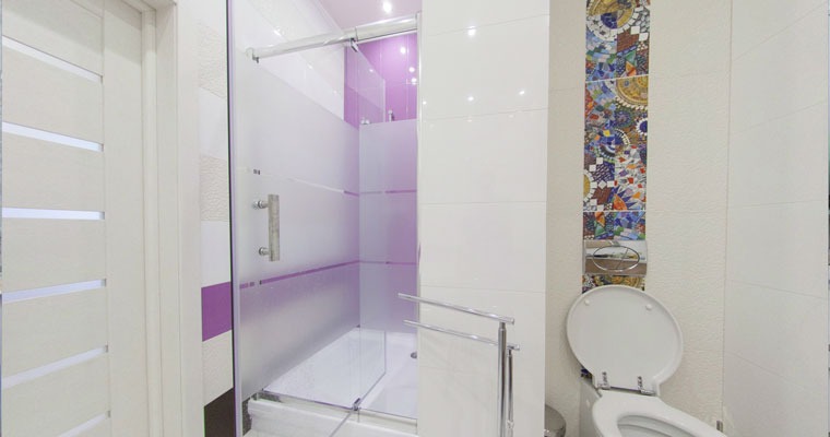 Nowoczesna łazienka z przeszkloną kabiną prysznicową, białą armaturą i mozaiką dekoracyjną na ścianie, widok na białe drzwi z poziomymi wgłębieniami.