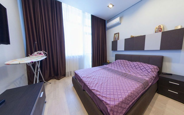 Sypialnia z łóżkiem z brązowym wezgłowiem i fioletową narzutą w geometryczny wzór, na ścianie wiszące szafki w szaro-brązowej kolorystyce, obok okna deska do prasowania