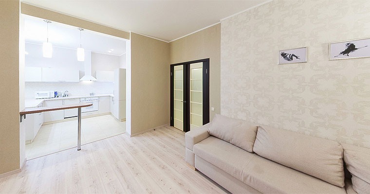 Jasne wnętrze mieszkania z aneksem kuchennym, beżowa sofa na pierwszym planie, na ścianie dekoracyjne tapety i dwa obrazy w srebrnych ramkach.