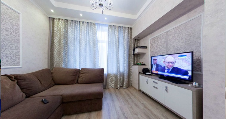 Jasne wnętrze salonu z brązową narożną sofą, wzorzystymi tapetami i zasłonami, oraz telewizorem na białej komodzie.