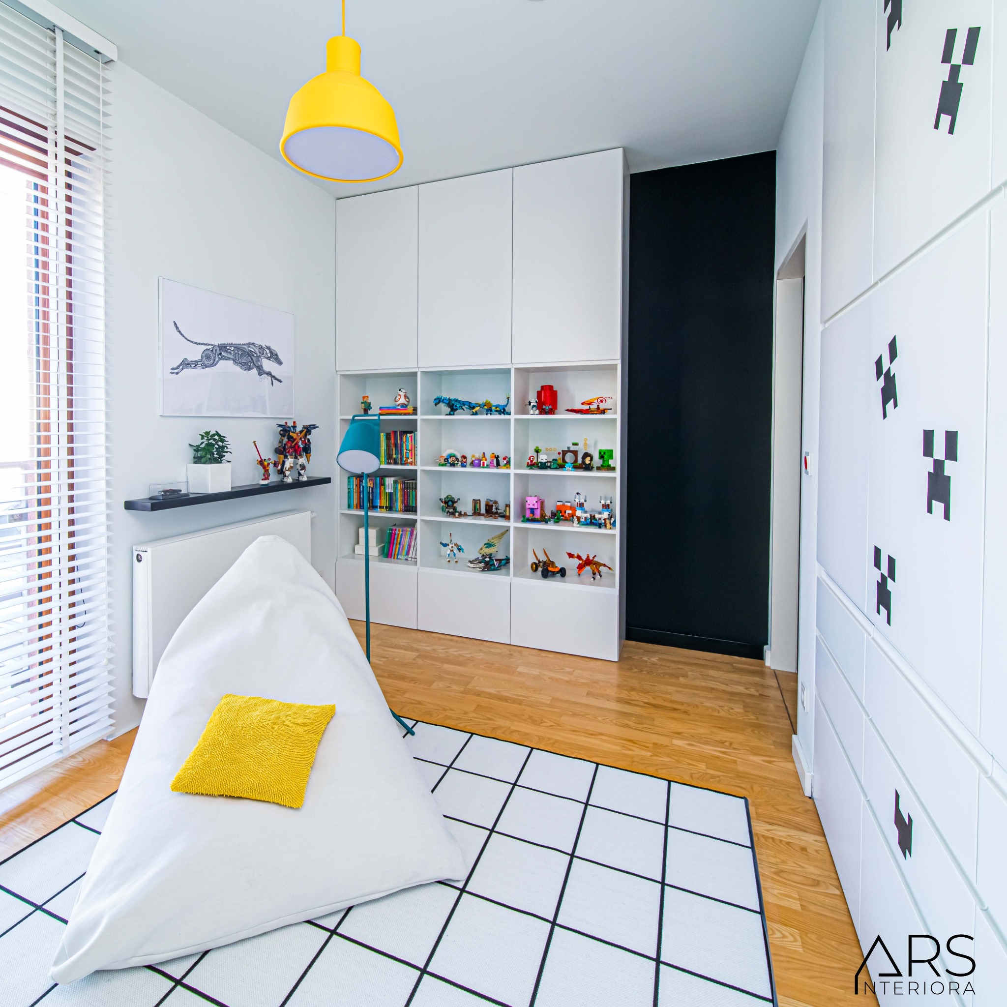 Pokój dziecięcy z białymi meblami, żółtą lampą i pufą w kształcie piramidy, dywan w czarno-białą kratę, dekoracje Minecraft na szafie, półki z zabawkami i książkami.