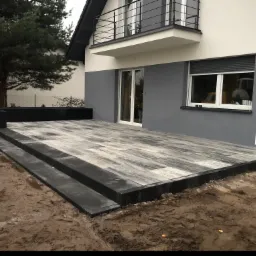 Nowo ułożony taras z szaro-białej kostki brukowej przed domem z balkonem, widoczne dwustopniowe schody z czarnej kostki i rabata w tle, ziemia wokół jeszcze niezagospodarowana.