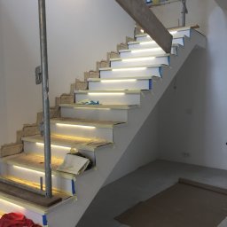 Bogdan - Montaż schodów z podświetleniem LED w trakcie wykańczania, widoczne zabezpieczenia taśmą malarską, surowe ściany i prowizoryczne podpory, ochrona podłogi kartonem.