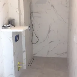 Nowoczesna łazienka w trakcie wykończenia, płytki imitujące marmur na ścianach i podłodze, widoczna kabina prysznicowa typu walk-in, narzędzia budowlane w rogu pomieszczenia.