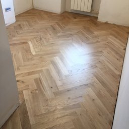 Bogdan - Jasny parkiet ułożony we wzór jodełki w pokoju z białymi ścianami i grzejnikiem, widok z poziomu podłogi.