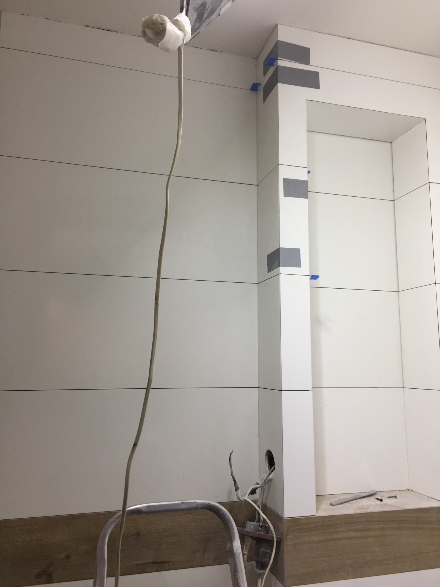Wnętrze łazienki w trakcie prac wykończeniowych, widoczne białe płytki imitujące panele drewniane, zabezpieczone narożniki, wystające kable i drabina.