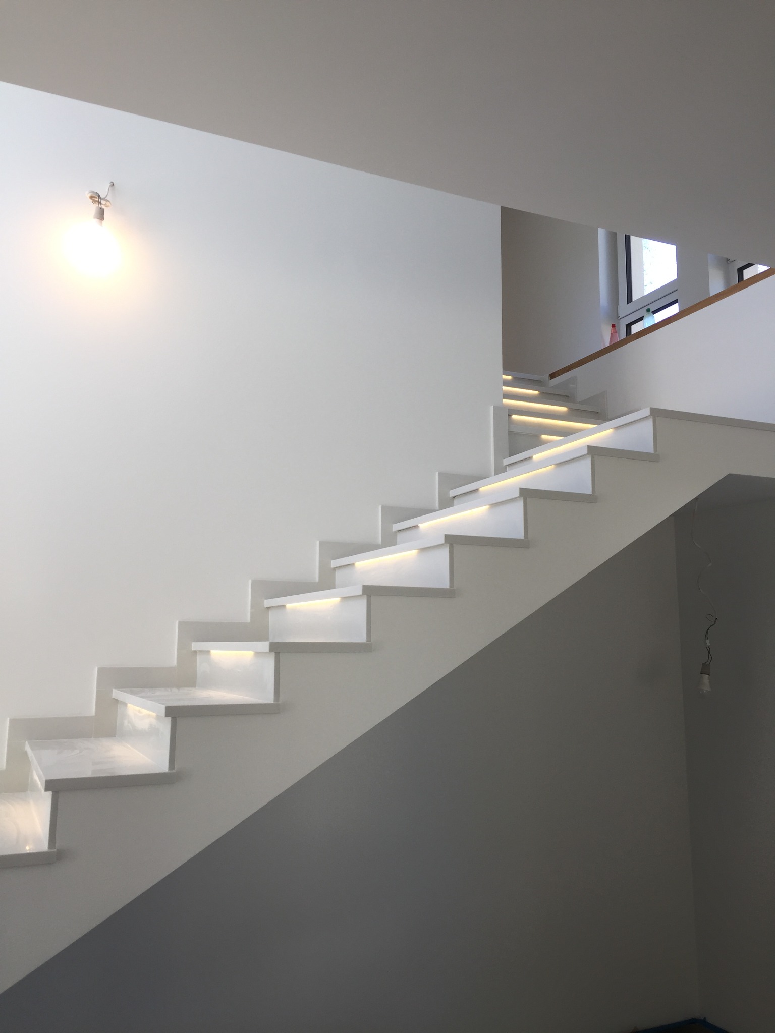 Nowoczesne, białe schody z podświetleniem LED pod stopniami, widoczne zawieszone kable elektryczne i żarówka na ścianie