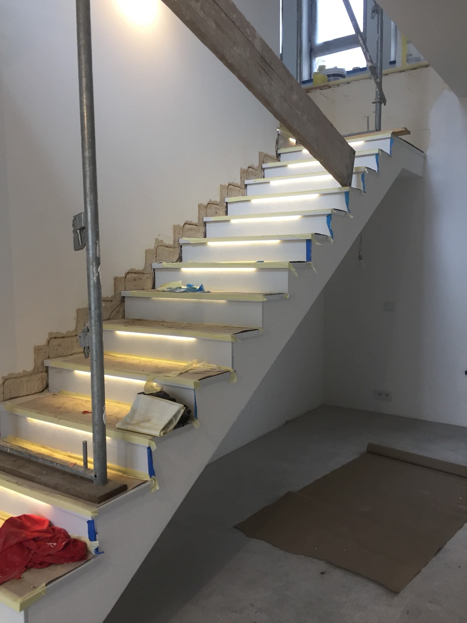 Montaż schodów z podświetleniem LED w trakcie wykańczania, widoczne zabezpieczenia taśmą malarską, surowe ściany i prowizoryczne podpory, ochrona podłogi kartonem.