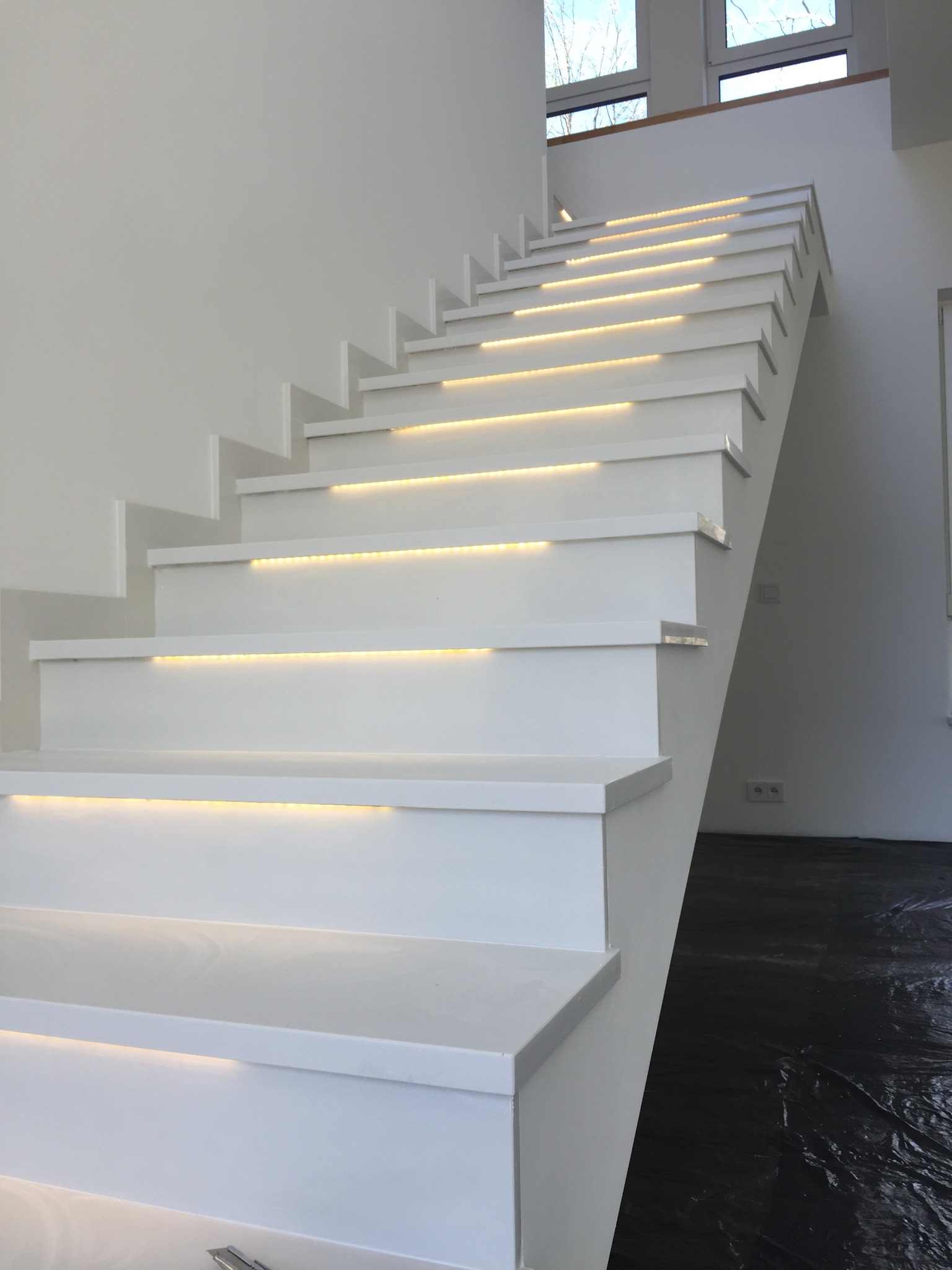 Nowoczesne, białe schody z podświetleniem LED na każdym stopniu, prowadzące do góry w jasnym wnętrzu z widocznymi oknami i zabezpieczoną folią podłogą.