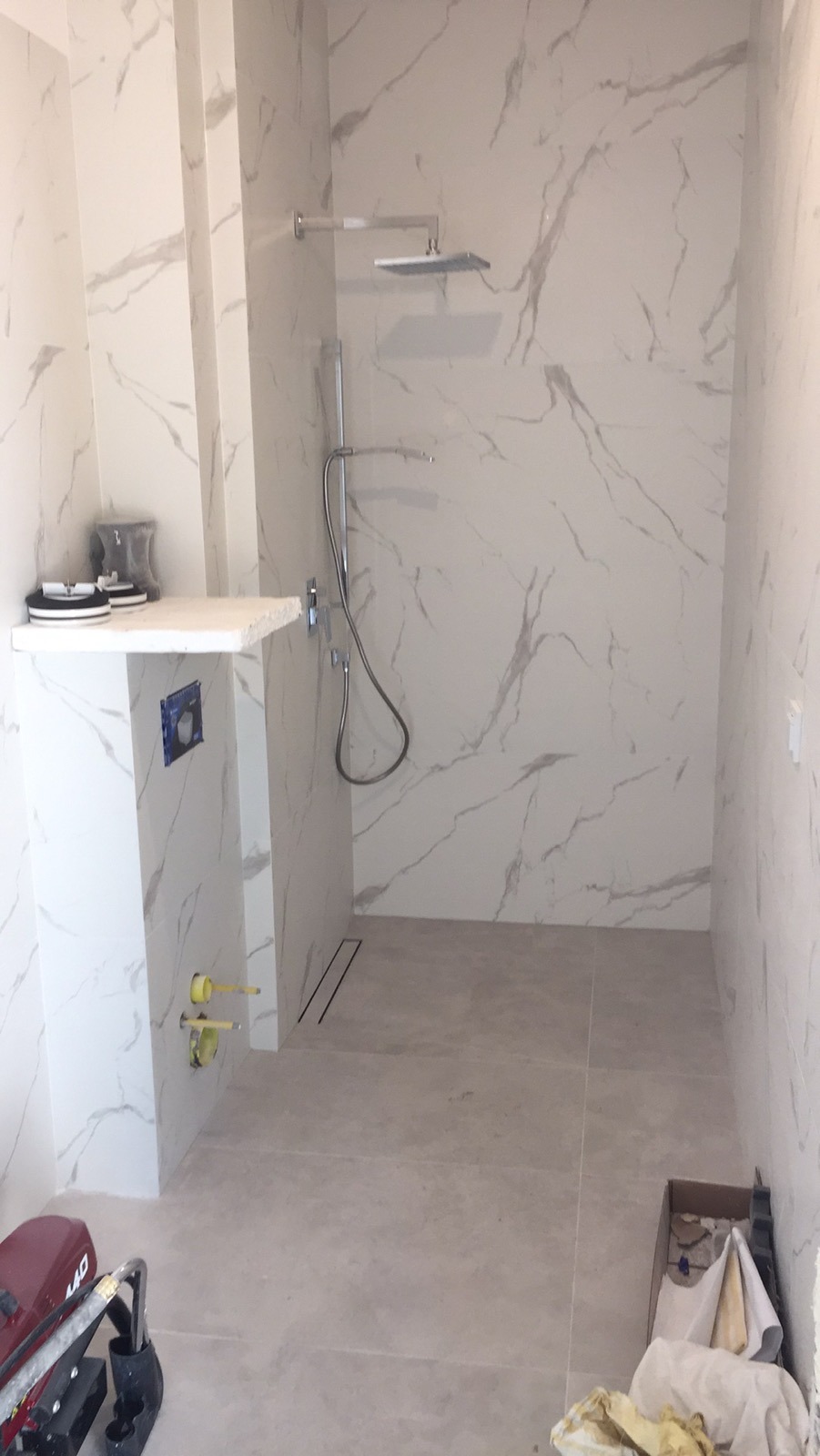 Nowoczesna łazienka w trakcie wykończenia, płytki imitujące marmur na ścianach i podłodze, widoczna kabina prysznicowa typu walk-in, narzędzia budowlane w rogu pomieszczenia.