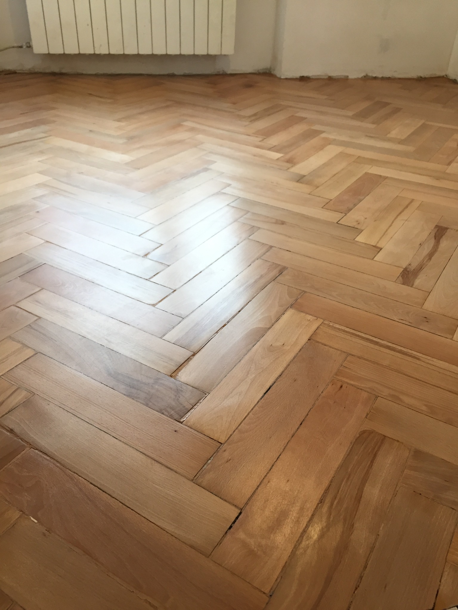 Parkiet ułożony w jodełkę francuską, widoczne ślady użytkowania i delikatny połysk, z fragmentem białego grzejnika żeberkowego w tle, przy ścianie wymagającej wykończenia.