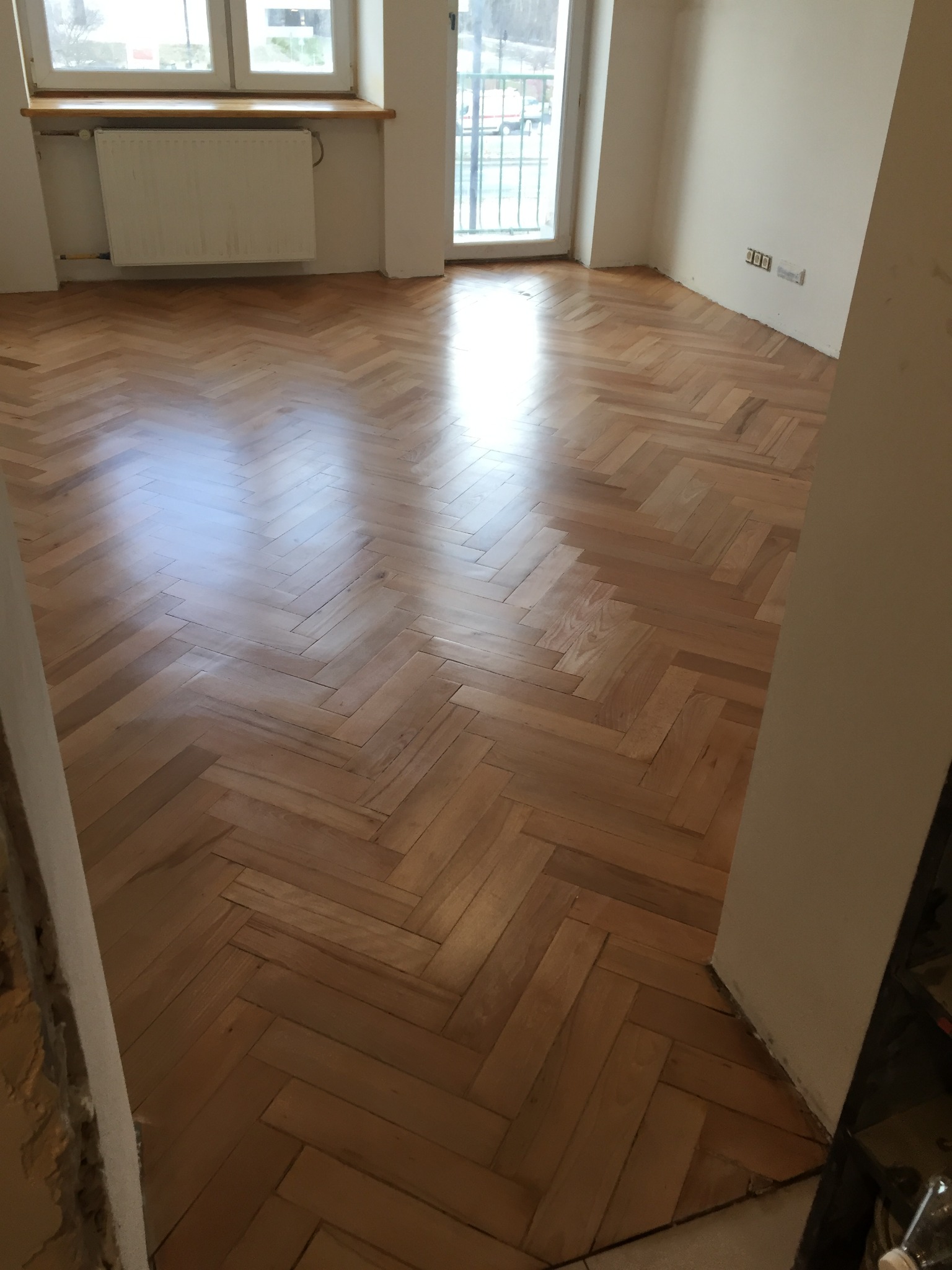 Wypolerowany parkiet ułożony w jodełkę w jasnym, pustym pokoju z oknem i grzejnikiem.