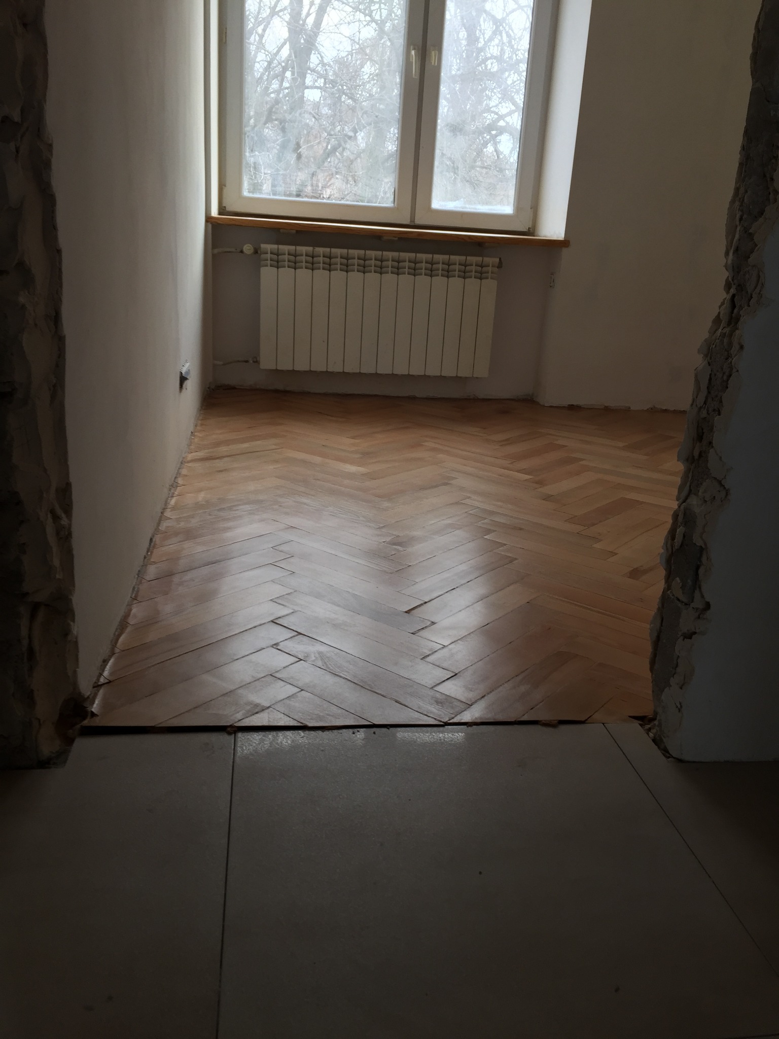 Ukończona podłoga z parkietu ułożonego w jodełkę w pokoju z grzejnikiem pod oknem, widoczne ślady po pracach remontowych na ścianach.
