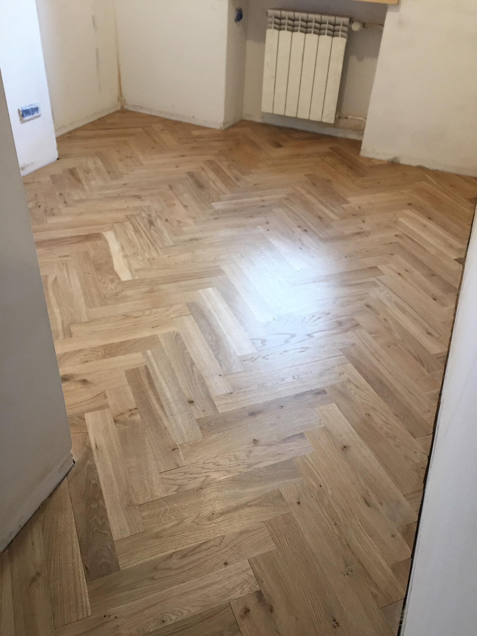 Jasny parkiet ułożony we wzór jodełki w pokoju z białymi ścianami i grzejnikiem, widok z poziomu podłogi.