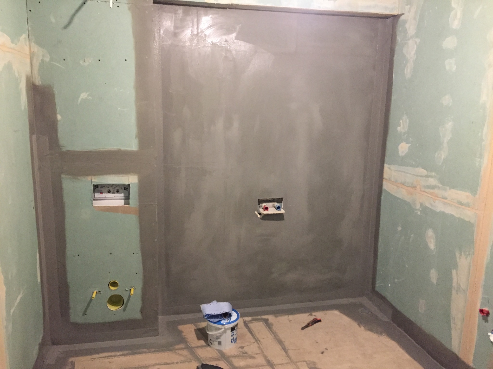 Przygotowanie ścian w łazience do położenia płytek: zagruntowane ściany, uszczelnione narożniki, widoczne otwory na instalacje wodno-kanalizacyjne, wiaderko z preparatem uszczelniającym na podłodze.