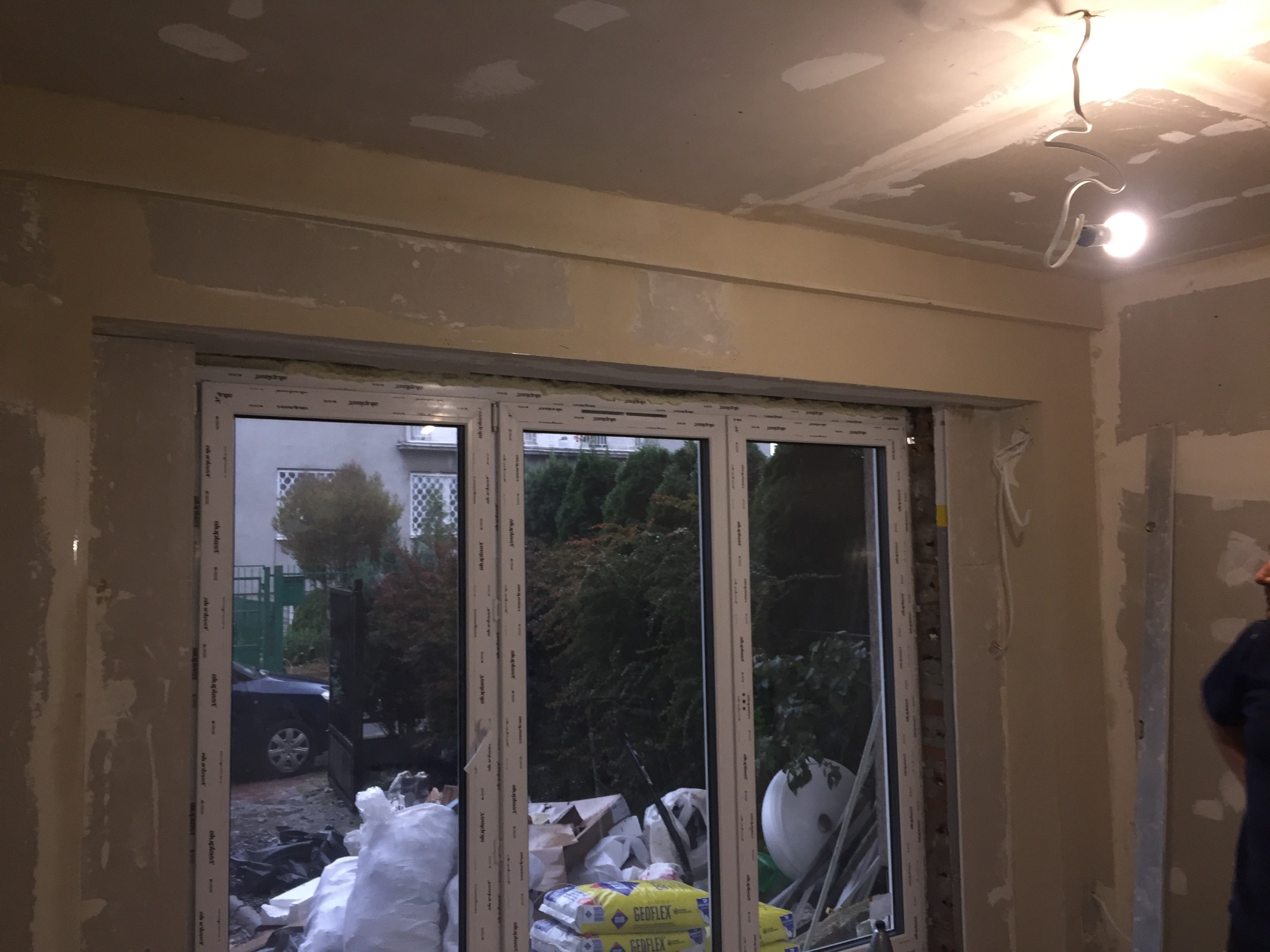 Wnętrze w trakcie prac wykończeniowych: zamontowane, trzyskrzydłowe okno z widokiem na podwórze, surowy sufit z widocznymi śladami szpachlowania, prowizoryczne oświetlenie i materiały budowlane...