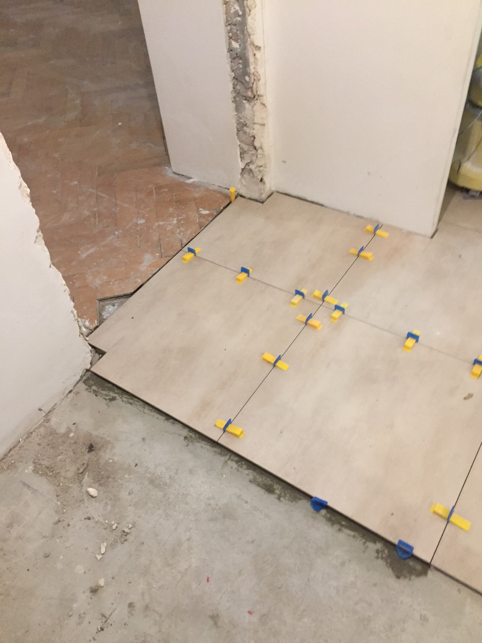 Układanie beżowych płytek podłogowych z użyciem żółto-niebieskich klipsów poziomujących, widoczny fragment starej, usuniętej podłogi, ślady zaprawy na surowym betonie.
