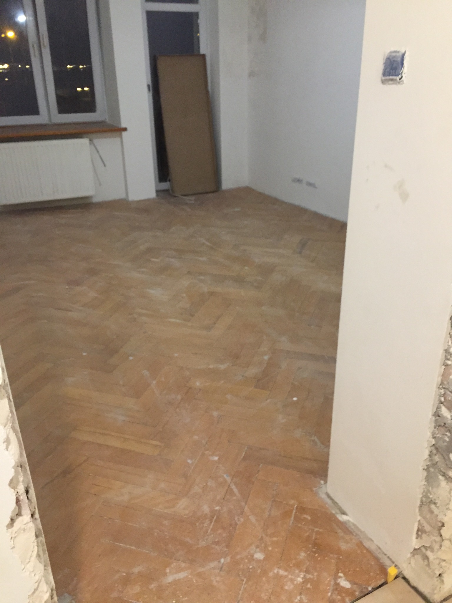 Pokój w trakcie remontu z parkietem ułożonym w jodełkę, ściany w stanie surowym, widoczne ślady po instalacjach elektrycznych, okno z widokiem na nocne miasto.