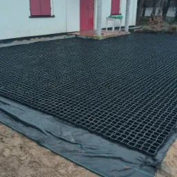 Przygotowany teren pod trawnik z siatką wzmacniającą i geowłókniną, widoczny fragment domu z czerwonymi okiennicami w tle.