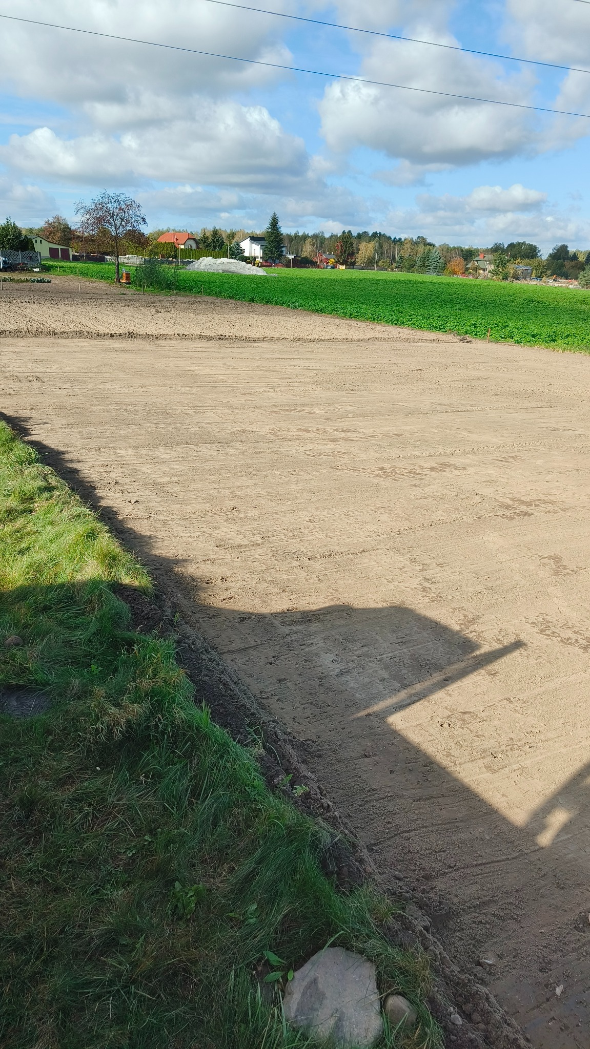 Przygotowany, wyrównany teren pod przyszły ogród, widoczne ślady po maszynach, w tle zielone pole i zabudowania.
