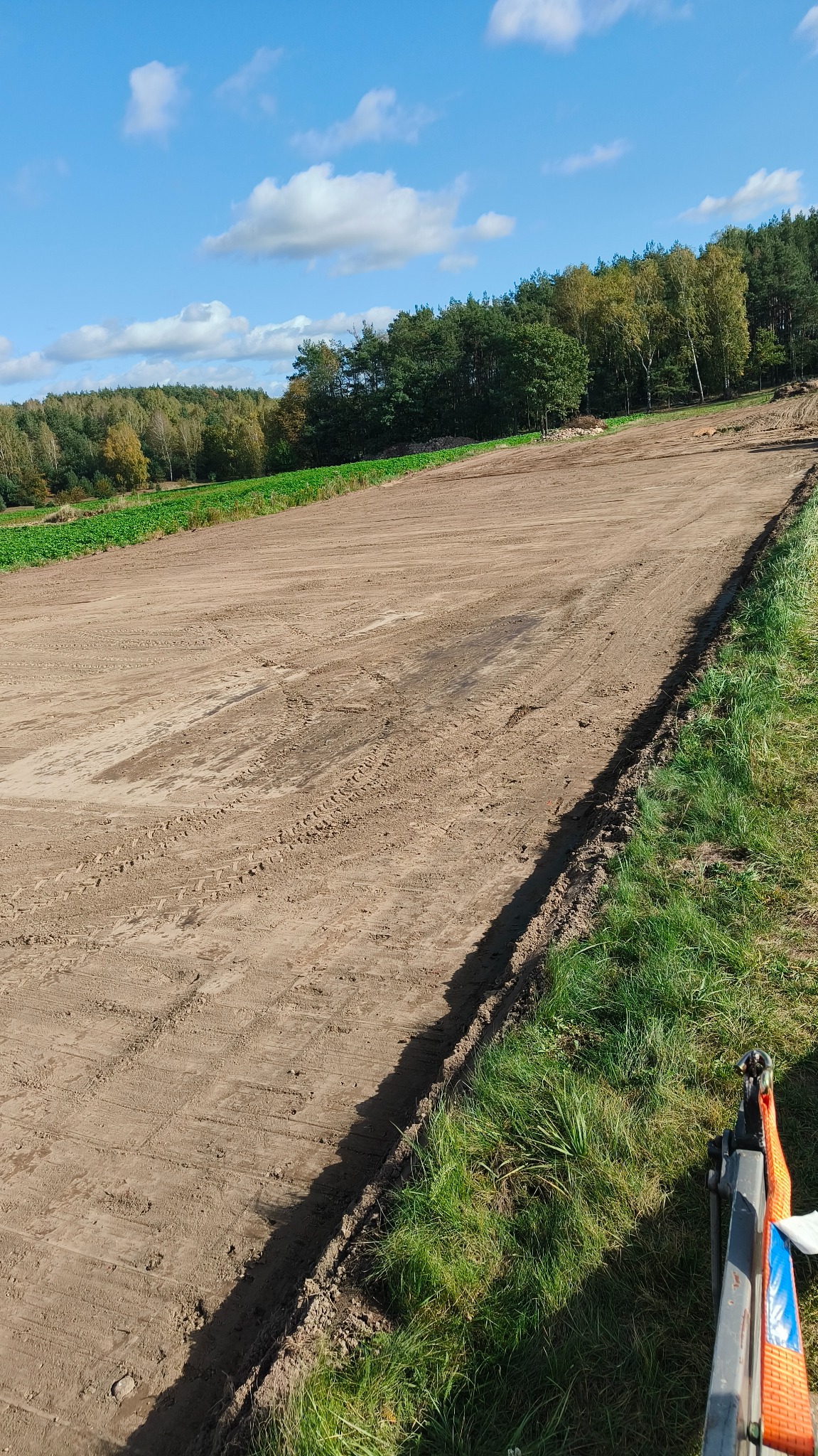 Przygotowany teren pod budowę lub ogród, wyrównana ziemia z widocznymi śladami opon, w tle las i błękitne niebo z chmurami.