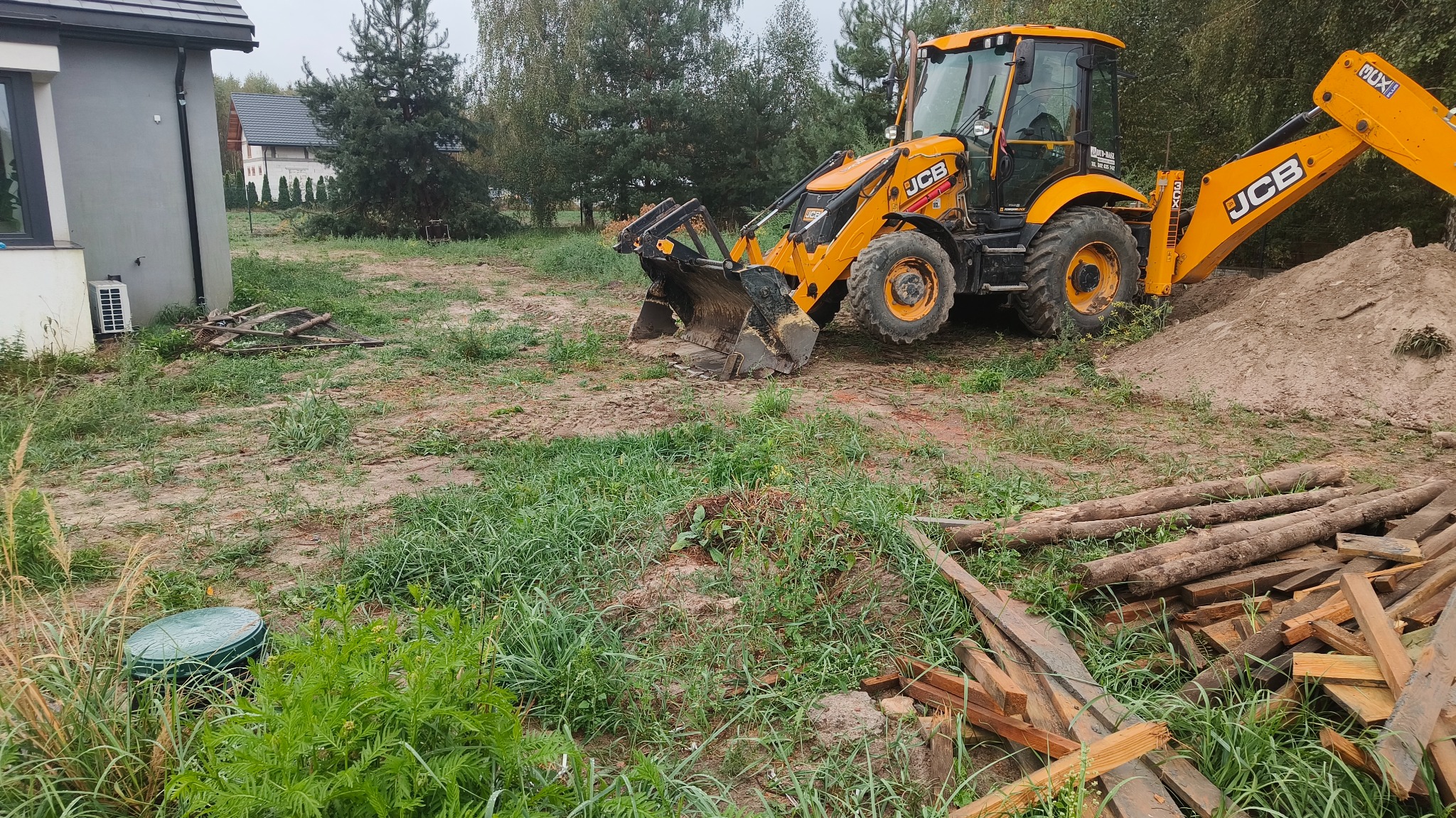 Żółta koparko-ładowarka JCB na działce z nierównym terenem, sterta ziemi obok, w tle dom i drzewa, widoczne ślady pracy maszyny.