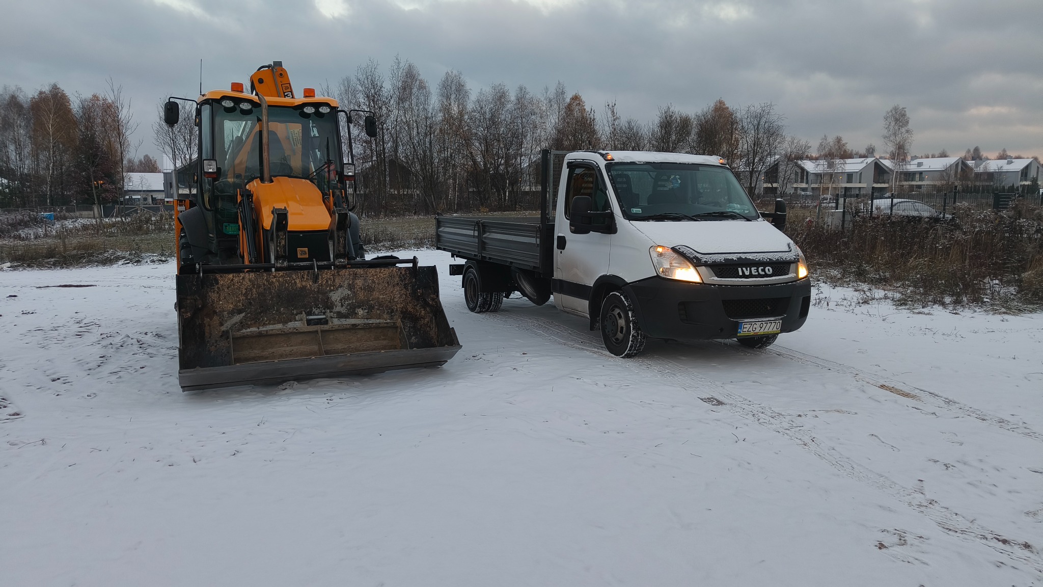 Pomarańczowa koparko-ładowarka JCB z brudną łyżką i biały samochód dostawczy Iveco na zasypanym śniegiem placu budowy w pochmurny dzień.