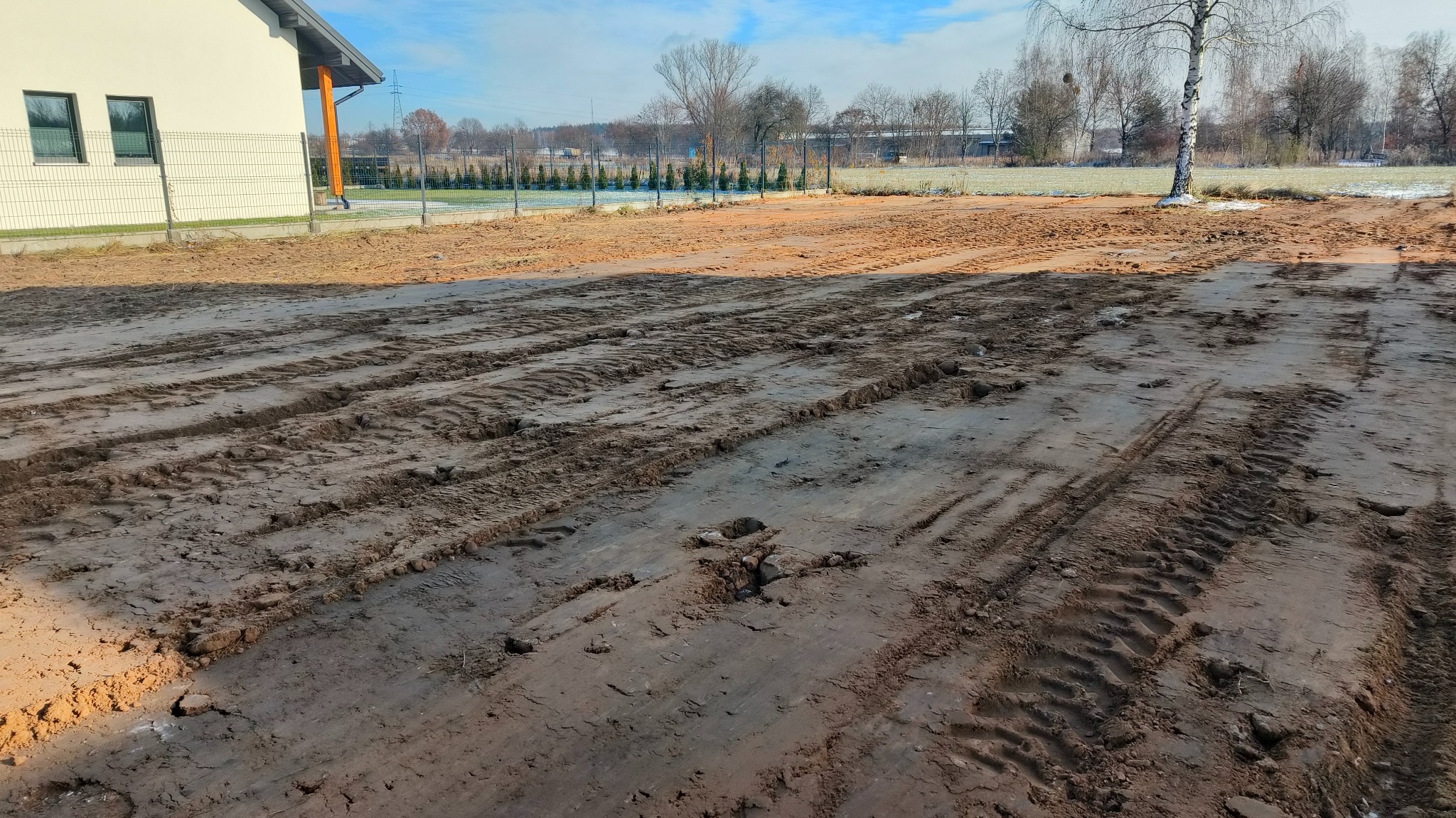 Przygotowany teren pod trawnik z widocznymi śladami opon, częściowo oświetlony słońcem, z budynkiem w tle.