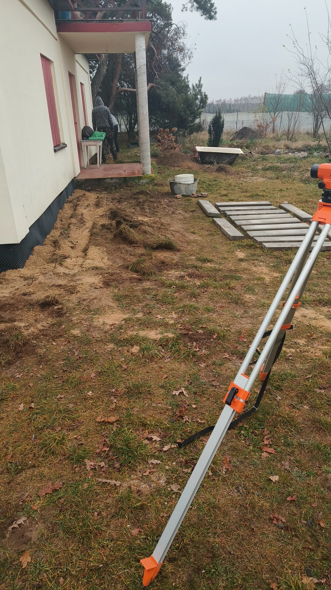 Niwelacja terenu przy domu jednorodzinnym za pomocą niwelatora optycznego na trójnogu, widoczne przygotowane płyty chodnikowe i pracownik w tle.