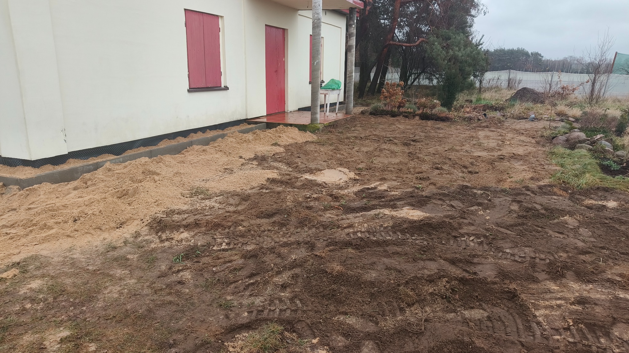 Przygotowanie terenu pod trawnik: warstwa piasku przy budynku z czerwonymi okiennicami, dalej teren z błotnistymi śladami opon.
