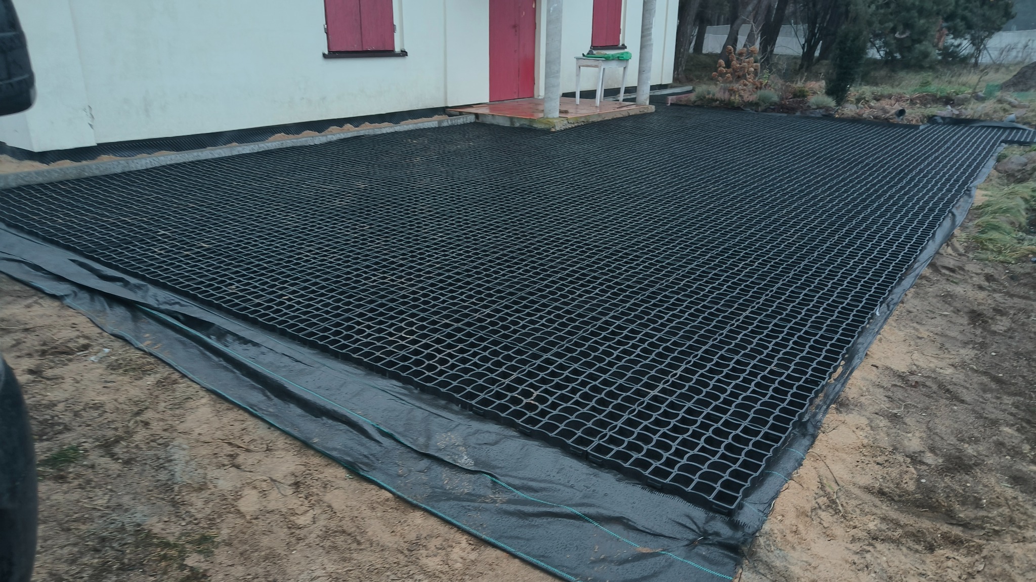 Przygotowany teren pod trawnik z siatką wzmacniającą i geowłókniną, widoczny fragment domu z czerwonymi okiennicami w tle.