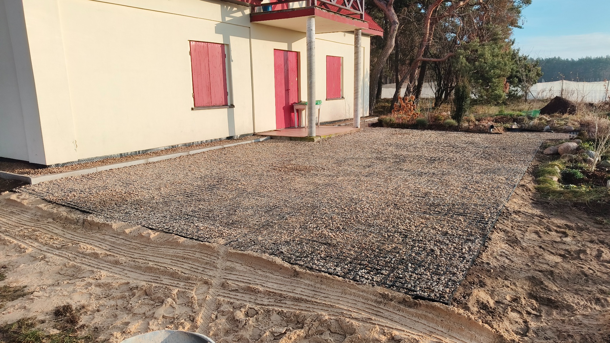 Przygotowany teren pod nawierzchnię z kruszywa, widoczna siatka wzmacniająca, obrzeża budynku z czerwonymi okiennicami i przygotowane podłoże z piasku.
