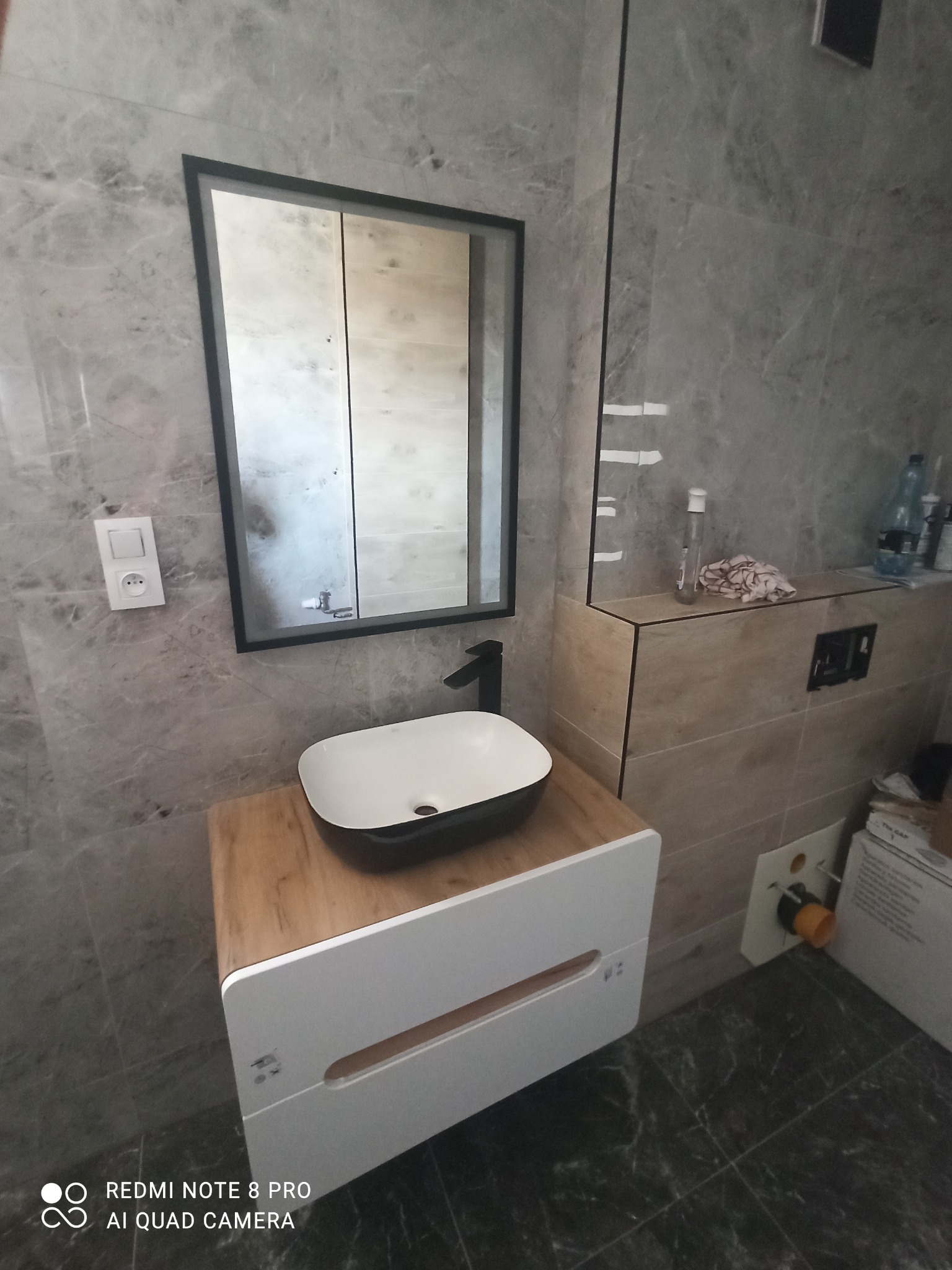 Nowoczesna łazienka z szarymi marmurowymi płytkami, umywalką nablatową na drewnianej szafce z białymi frontami, lustrem w czarnej ramie i widoczną instalacją hydrauliczną.