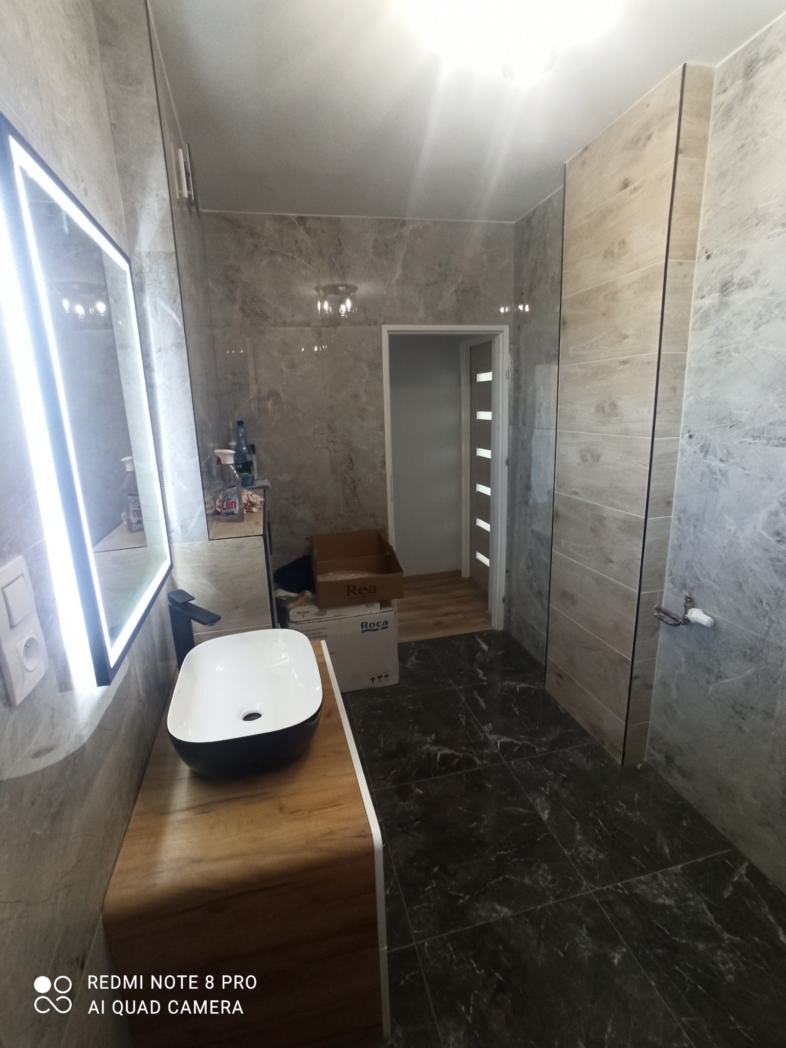 Nowoczesna łazienka z marmurowymi płytkami, podwieszaną umywalką w czarno-białej kolorystyce i lustrem z podświetleniem LED, widoczne kartony po armaturze.