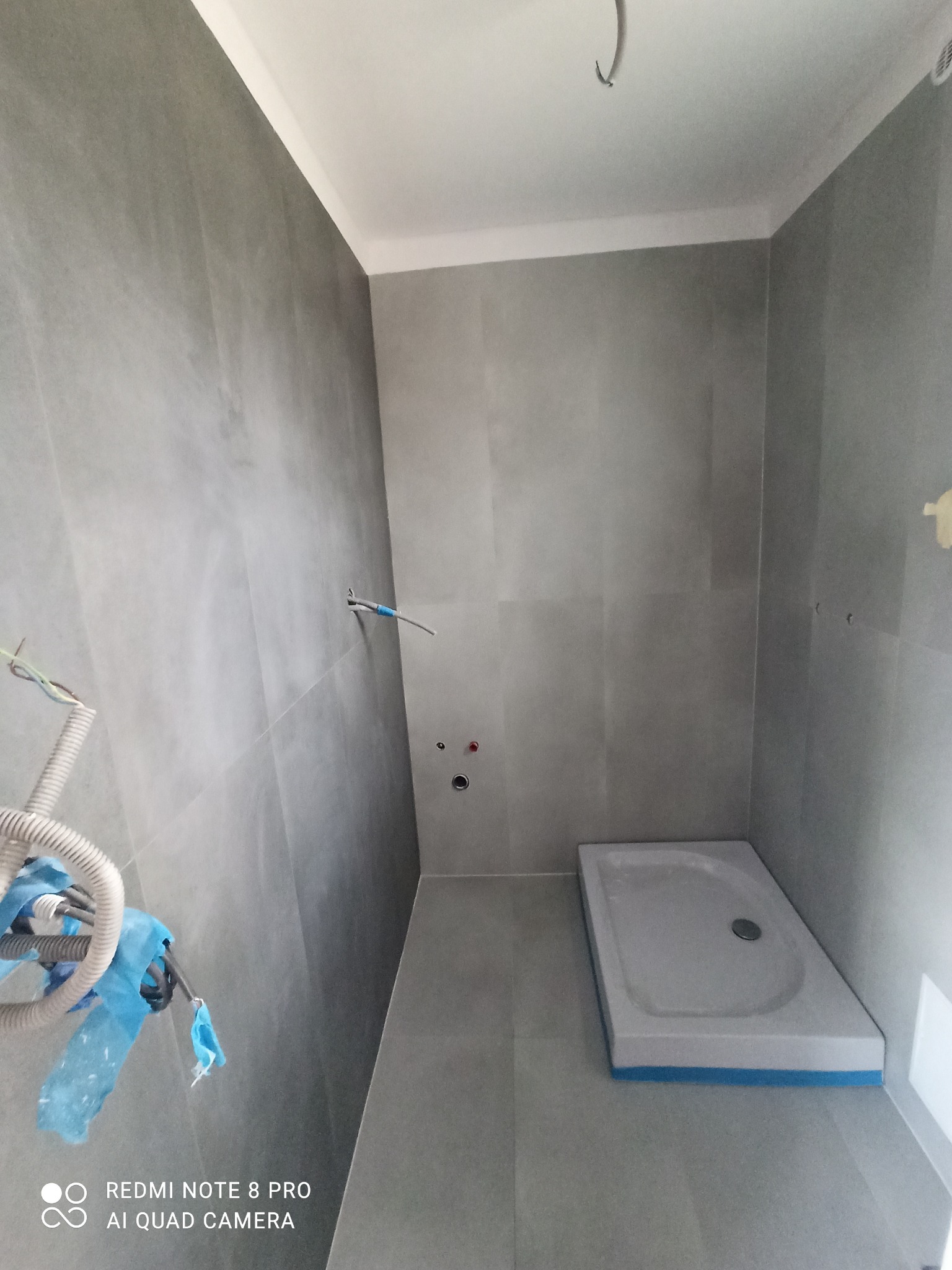 Płytki imitujące beton na ścianach i podłodze w surowej łazience, widoczny brodzik, wystające kable i rury przed podłączeniem armatury.