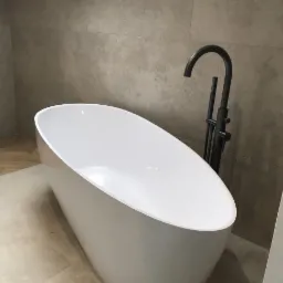 Nowoczesna, wolnostojąca wanna owalna z czarną armaturą na tle płytek w odcieniach beżu i szarości, oświetlona światłem dziennym z okna dachowego.