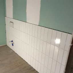 Ściana w trakcie układania białych, prostokątnych płytek, widoczne niebieskie zawory i fragment podłogi z paneli imitujących drewno oraz fragment ściany pokrytej zieloną farbą i pasami gipsu.