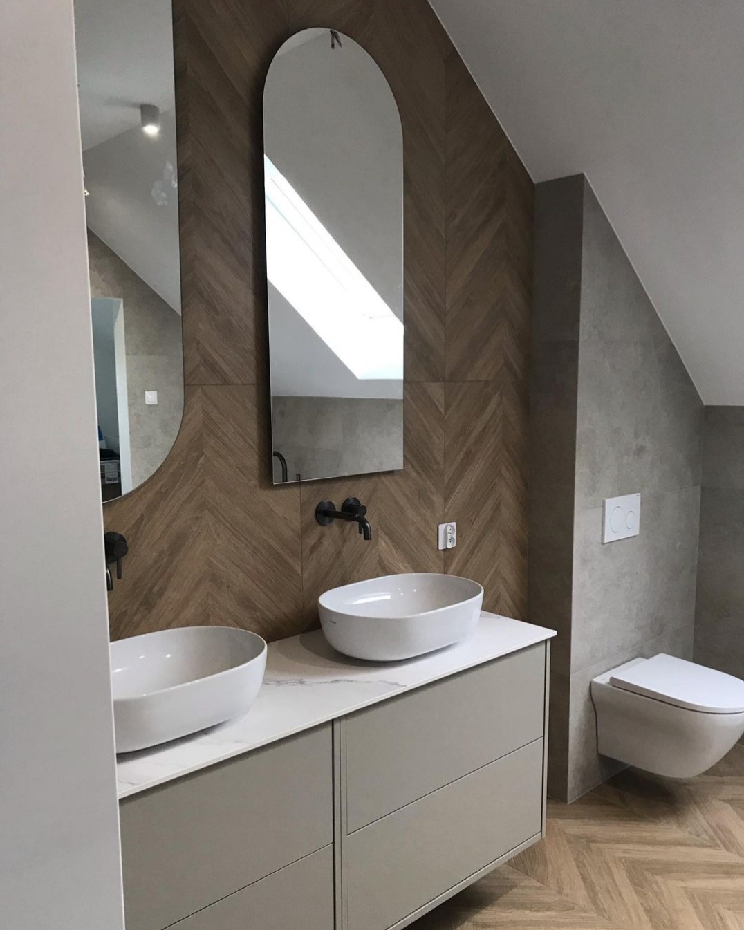 Nowoczesna łazienka na poddaszu z dwoma owalnymi umywalkami nablatowymi, lustrami o zaokrąglonym kształcie i drewnopodobną glazurą ułożoną w jodełkę.