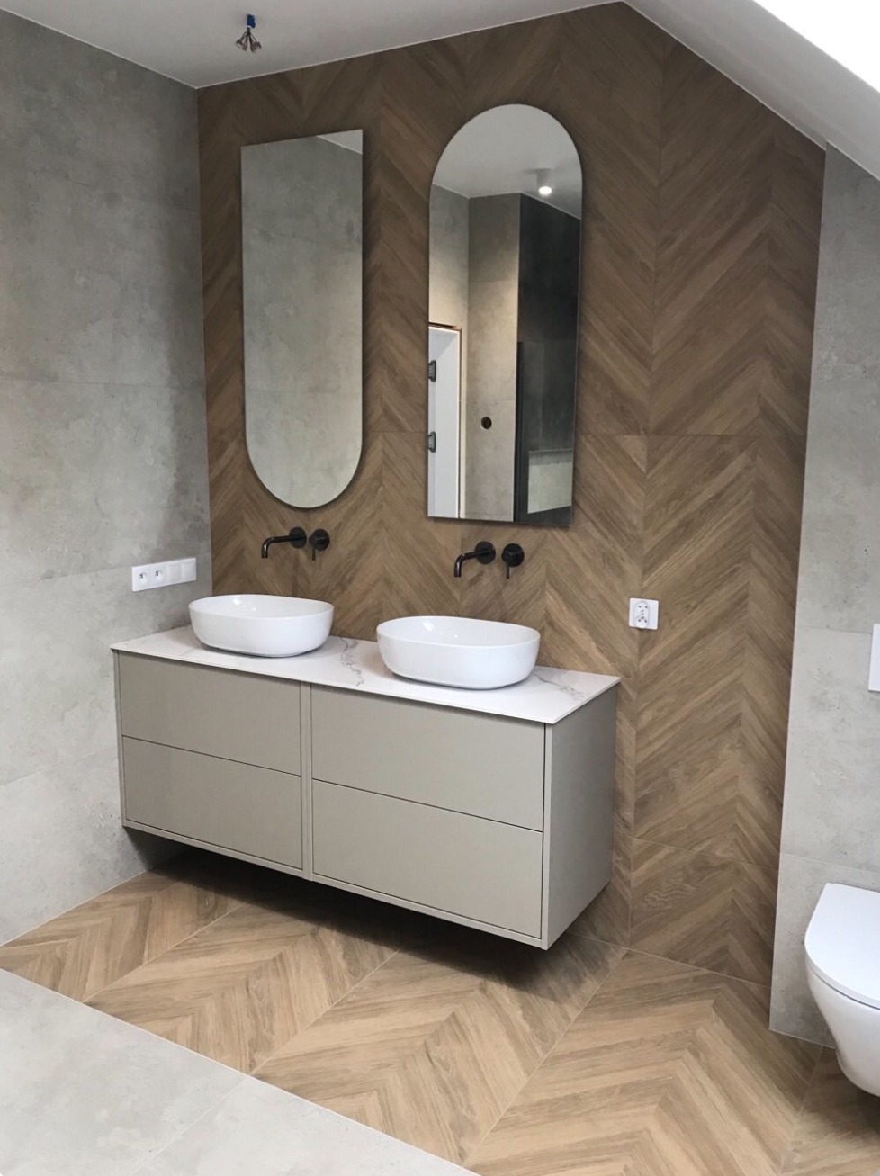 Nowoczesna łazienka z dwoma owalnymi umywalkami nablatowymi, szafką podumywalkową w odcieniach szarości, dwoma podłużnymi lustrami z zaokrąglonymi narożnikami oraz ścianą wyłożoną płytkami...