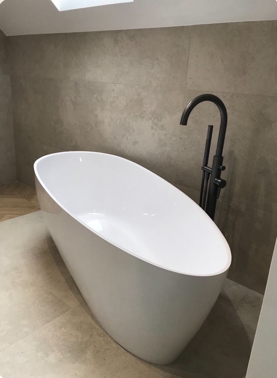 Nowoczesna, wolnostojąca wanna owalna z czarną armaturą na tle płytek w odcieniach beżu i szarości, oświetlona światłem dziennym z okna dachowego.