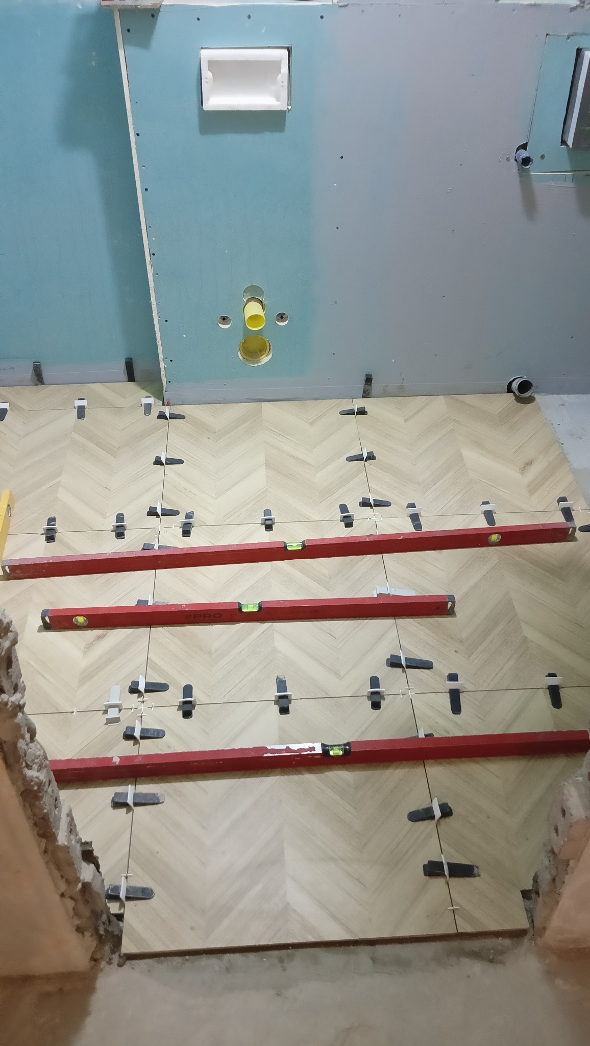 Układanie parkietu ceramicznego w jodełkę w trakcie remontu łazienki, z użyciem poziomnic i klipsów dystansowych, widoczne przygotowane ściany z instalacją podtynkową.