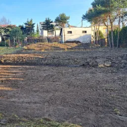 Przygotowany teren pod budowę z wykopem i wyrównanym gruntem, widoczne ślady po maszynach budowlanych, w tle budynek mieszkalny i drzewa.