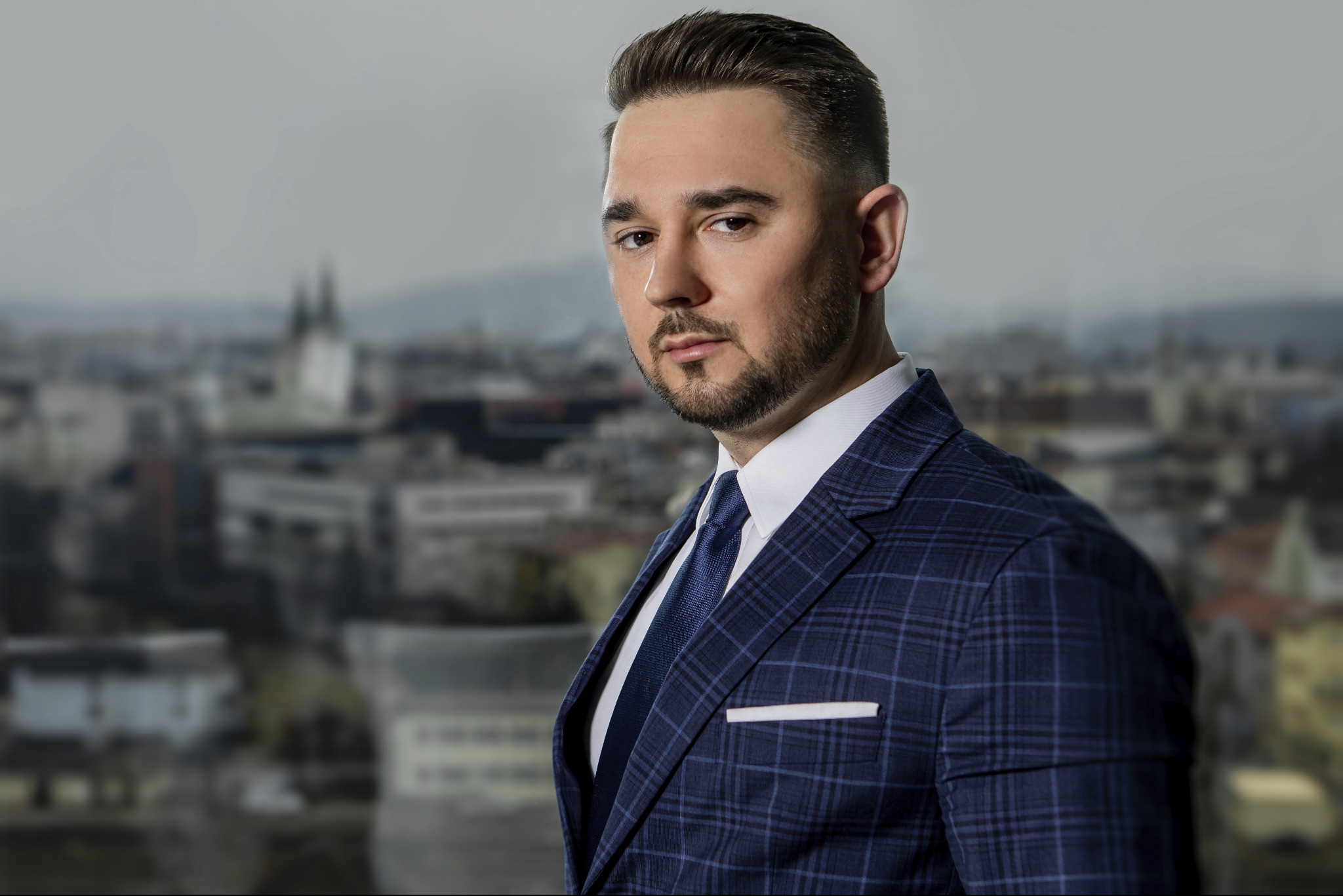 Portret mężczyzny w niebieskiej kraciastej marynarce i krawacie na rozmytym tle miejskiego krajobrazu.