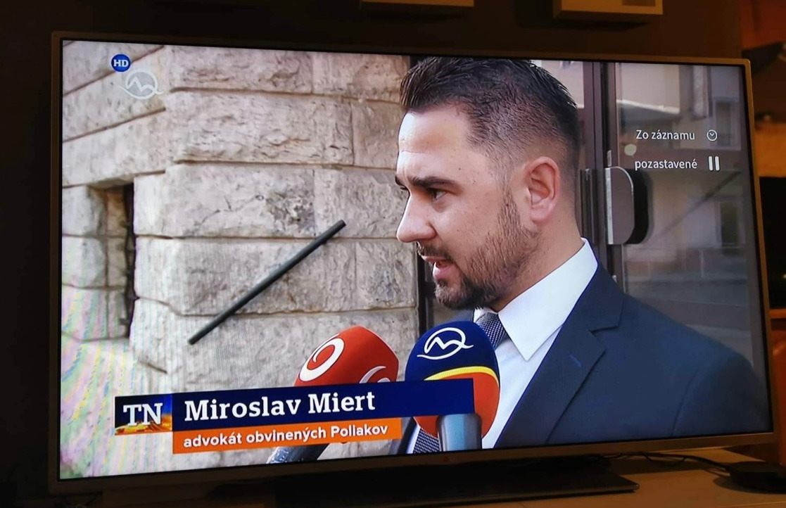 Mężczyzna w garniturze udziela wywiadu przed budynkiem, widoczny pasek informacyjny z nazwiskiem i tytułem 'adwokat obviněných Pollakov'.